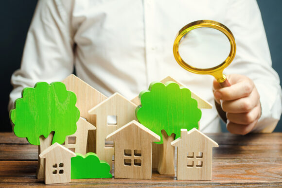 A man holds a magnifying glass over wooden houses and trees. Real estate valuation and selection of a place for the construction of new buildings. Evaluation of urbanism and residential space.